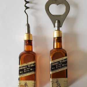 May include: Two vintage Johnnie Walker Old Scotch Whisky bottle-shaped bar tools. One is a corkscrew, the other a bottle opener. Both have amber-colored glass bodies, gold-colored metal accents, and labels with text and a walking figure.