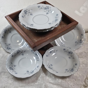 May include: A set of white porcelain saucers and bowls with a delicate blue floral pattern and silver trim. The bowls are stacked on a wooden tray, with additional saucers arranged around it. The set is ideal for serving tea or desserts.