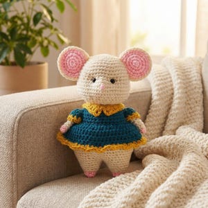 Pode incluir: Boneca de rato de crochê com corpo creme, orelhas grandes rosa e creme e olhos pretos. Ela usa um vestido azul-petróleo com gola e acabamento amarelos. A boneca tem pés e mãos rosa e está sentada em um sofá bege.