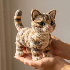 Op de afbeelding: Een handgemaakte gehaakte kattenfiguur in tinten bruin, beige en crème. De kat heeft groene ogen, een roze neus en een gestreepte staart. De kat staat rechtop, met zijn poten zichtbaar. Het is een decoratief item.