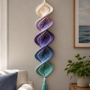 Può includere: Un arazzo verticale all'uncinetto con un design a spirale e una sfumatura di colore che passa dal crema in alto al viola, blu e turchese in basso. Il pezzo è rifinito con una perlina di legno e una nappa turchese abbinata.