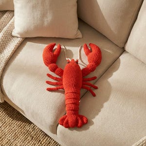 Puede incluir: Un cojín decorativo de langosta roja brillante, tejido a ganchillo, descansa sobre un sofá beige. La langosta tiene pinzas, patas y cola detalladas. Esta pieza es un acento caprichoso que añade un toque de encanto costero al espacio.