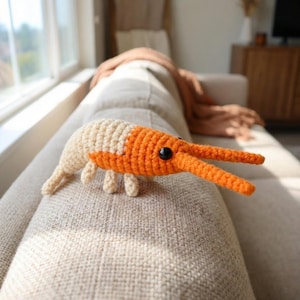 May include: A handmade crochet shrimp toy in cream and orange. The shrimp has black eyes and long orange pincers. The toy is resting on a beige sofa. This whimsical toy is perfect for children or as a decorative item.