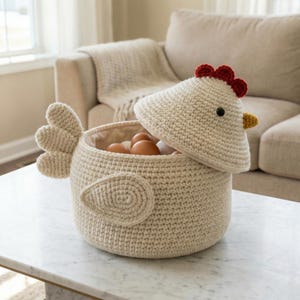 May include: A cream-colored crochet chicken-shaped egg basket with a removable lid. The lid features a red comb, a black eye, and a yellow beak. The basket is filled with brown eggs and sits on a white marble table.