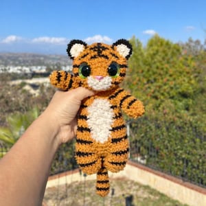 May include: A handmade orange and black striped tiger plush toy with large green eyes, a white belly, and a pink nose. The tiger has small black ears and is being held up against a background of trees and a blue sky.