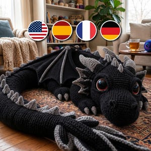 Pode incluir: Um grande peluche de dragão feito à mão em croché, com um corpo preto texturizado e espigões cinzentos claros contrastantes ao longo das costas, cauda e cabeça. A criatura tem olhos escuros brilhantes com íris castanho-avermelhadas.
