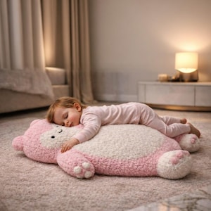 Puede incluir: Un niño dormido descansa sobre un gran cojín de suelo con forma de oso rosa y blanco. El cojín tiene una superficie suave y texturizada y presenta una cara y patas de oso. El niño lleva un pijama rosa.