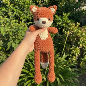 Puede incluir: Un peluche de zorro tejido a mano en tonos marrón, crema y blanco. El zorro tiene una textura esponjosa, con un pecho color crema y una cola con punta blanca. El juguete se sostiene sobre un fondo de follaje verde.