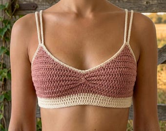 May include: A handmade crochet top in a dusty rose color with a cream-colored trim and straps. The top features a V-neck design and a textured stitch pattern, showcasing a bohemian style.
