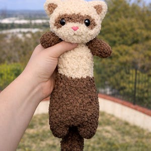 May include: A handmade ferret plush toy with a cream-colored head and body, and brown accents. The ferret has black eyes and a pink nose. The plush is held up against a blurred outdoor background.