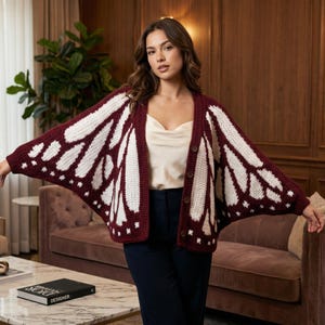 May include: A burgundy and white butterfly wing-patterned cardigan. The open-front sweater has a button closure and is made of a soft, textured material. The design features white wing patterns on a burgundy background. The cardigan is part of a clothing collection.