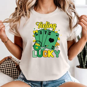 May include: Cream-colored t-shirt with a St. Patrick's Day graphic. The design features green playing cards with the words "LOVE" and a heart, a pot of gold, and the words "Feeling Lucky" in yellow and green.