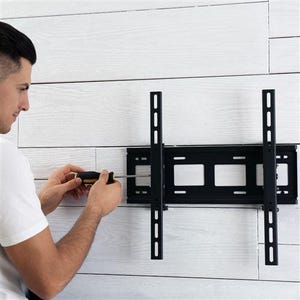 Puede incluir: Un soporte de pared para TV de metal negro se est&aacute; instalando en una pared de madera blanca. El soporte tiene brazos de soporte verticales y horizontales. Una persona est&aacute; usando un destornillador para asegurar el soporte.