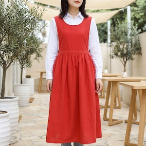 以下が含まれることがあります： 丸いネックラインとギャザーのスカートが付いた赤いエプロンドレス。エプロンは白い襟付きシャツの上に着用されています。ドレスは膝丈で、無地の赤い生地で作られています。背景には木、テーブル、石畳のパティオが含まれています。