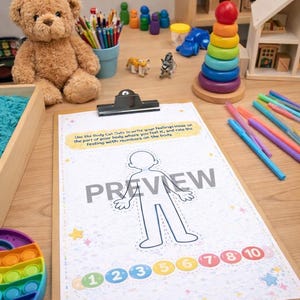 Puede incluir: Una mesa de madera exhibe una variedad de juguetes y herramientas para la terapia de juego. Se ven un oso de peluche, un juguete apilable de arcoíris y una hoja con un contorno corporal y números. El texto "Play Therapy" está encima de la mesa.