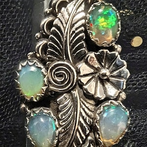 Puede incluir: Un anillo de plata con un diseño ornamentado que presenta cuatro ópalos ovalados y un motivo central de flor y hoja. Los ópalos muestran colores iridiscentes de verde, azul y amarillo. El anillo está colocado en un medidor de anillos.