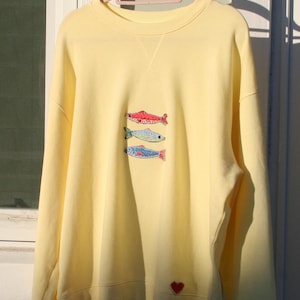 Puede incluir: Una sudadera amarilla pálida con peces bordados en rojo, azul y verde. Un pequeño corazón rojo está bordado cerca del dobladillo inferior. La sudadera está colgada en una percha rosa.