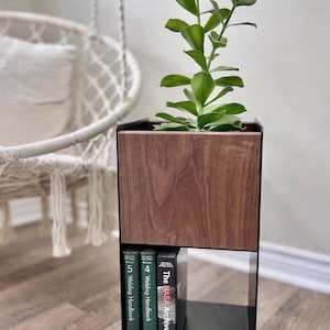 Puede incluir: Una jardinera moderna con una parte superior de vetas de madera oscura y un marco de metal negro. La jardinera contiene una planta verde frondosa y tres libros. Los libros se titulan "Welding Handbook" y "The NASA Archives".