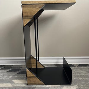 May include: A contemporary side table featuring a black metal frame and a wooden top and accents. The table has a distinctive geometric design with a shelf at the base. The wood displays a natural grain pattern.