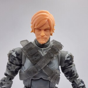 May include: A detailed action figure of a soldier with a light orange-colored head and beard. The figure wears a gray and black camouflage uniform with a tactical vest and shoulder pads. The figure's hair is styled in a short, layered cut.