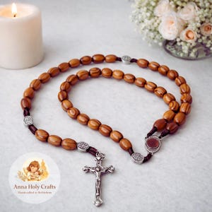 May include: A wooden rosary with oval beads and a silver crucifix. The rosary features a brown cord and silver accents. The image includes a lit candle, flowers, and a logo that reads "Anna Holy Crafts, Handcrafted in Bethlehem."