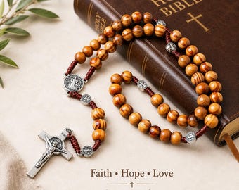 May include: A rosary with polished brown wooden beads strung on a dark red cord, featuring silver-toned metal medals and a crucifix with an INRI inscription. The rosary rests on a brown Holy Bible with the text Faith • Hope • Love printed below.