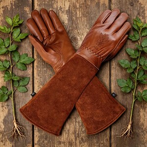 May include: Brown leather gardening gloves with long suede cuffs, crossed on a wooden surface. The gloves have a leaf design and zig-zag stitching. Green plants with roots are on either side of the gloves.