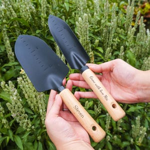 Könnte beinhalten: Zwei schwarze Gartenschaufeln mit Holzgriffen. Auf einem Griff steht "Steven", auf dem anderen "Grow with love Mia". Die Schaufeln haben Messmarkierungen auf der Klinge und ein Loch am Ende des Griffs.