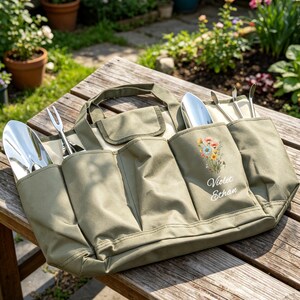 Puede incluir: Una bolsa verde de lona para herramientas de jard&iacute;n con m&uacute;ltiples bolsillos, que contiene varias herramientas de jardiner&iacute;a de acero inoxidable. La bolsa tiene un asa y un bordado floral con los nombres "Violet Ethan". La bolsa est&aacute; sobre una superficie de madera.