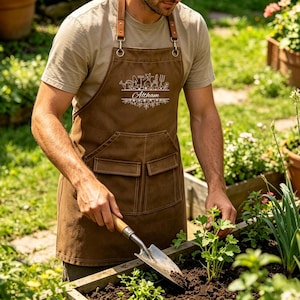 May include: A brown gardening apron with leather straps and pockets. The apron features a white floral design and the name "Altham". The person in the image is using a trowel to tend to plants in a garden bed.