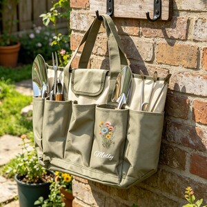 May include: An olive green and cream canvas garden tote bag, filled with gardening tools. The bag features multiple pockets and a floral design with the name "Molly" embroidered on one pocket. The bag is suspended on a brick wall.