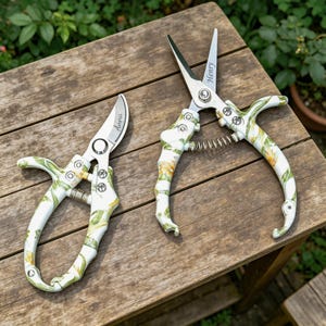May include: Two floral-patterned gardening shears with silver blades and white handles. One shear has "Anna" engraved on the blade, and the other has "Henry". The handles have a white base with green and yellow floral designs. The shears are on a wooden surface.