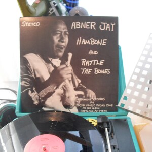 May include: A vinyl record album cover titled Abner Jay Hambone and Rattle the Bones, featuring a black and white portrait of a musician. The record is placed on a teal turntable with a black vinyl disc visible, alongside headphones and a metal stand.