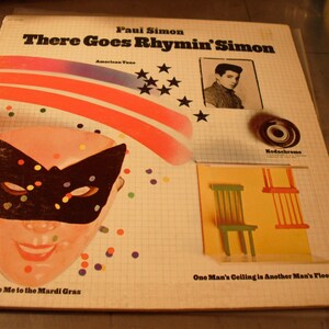 May include: Album cover for Paul Simon's "There Goes Rhymin' Simon." The cover features a portrait of Paul Simon, a Mardi Gras mask, and a colorful design with the text "American Tune." The album is in a protective sleeve.