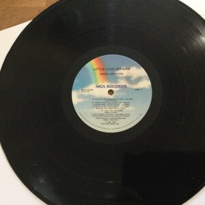 May include: A black vinyl record with the album title "Little Love Affairs" by Nanci Griffith. The record label features a rainbow, clouds, and the MCA Records logo. The record is in a white sleeve.