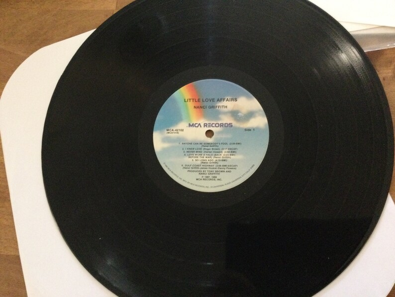 May include: A black vinyl record with the album title "Little Love Affairs" by Nanci Griffith. The record label features a rainbow, clouds, and the MCA Records logo. The record is in a white sleeve.