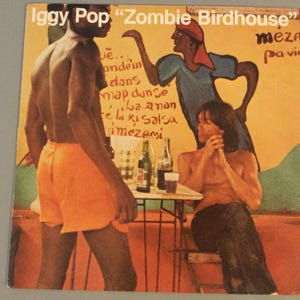 May include: Album cover for Iggy Pop's "Zombie Birdhouse" featuring three figures in a colorful, stylized illustration. The album art has text at the top and on the side. The background is a warm yellow with a mural-like quality.