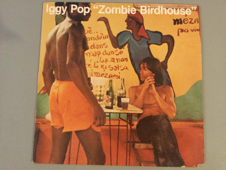 May include: Album cover for Iggy Pop's "Zombie Birdhouse" featuring three figures in a colorful, stylized illustration. The album art has text at the top and on the side. The background is a warm yellow with a mural-like quality.