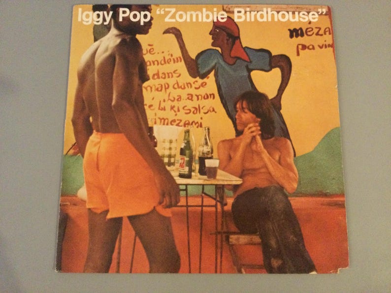 May include: Album cover for Iggy Pop's "Zombie Birdhouse" featuring a colorful illustration. The artwork depicts three figures, one in orange shorts, another in a blue and red outfit, and a third seated at a table. The background is a mix of orange, yellow, and green.