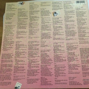 May include: The back cover of a vinyl record album with song lyrics printed in black on a pink and white background. The album's title and artist are not visible. The lyrics are arranged in columns, with a small photo in the upper left corner.