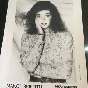 May include: Black and white promotional photo of Nanci Griffith. She is wearing a floral print blouse and a belt. Her dark hair is styled with volume. The photo includes the text "NANCI GRIFFITH" and "MCA RECORDS NASHVILLE".