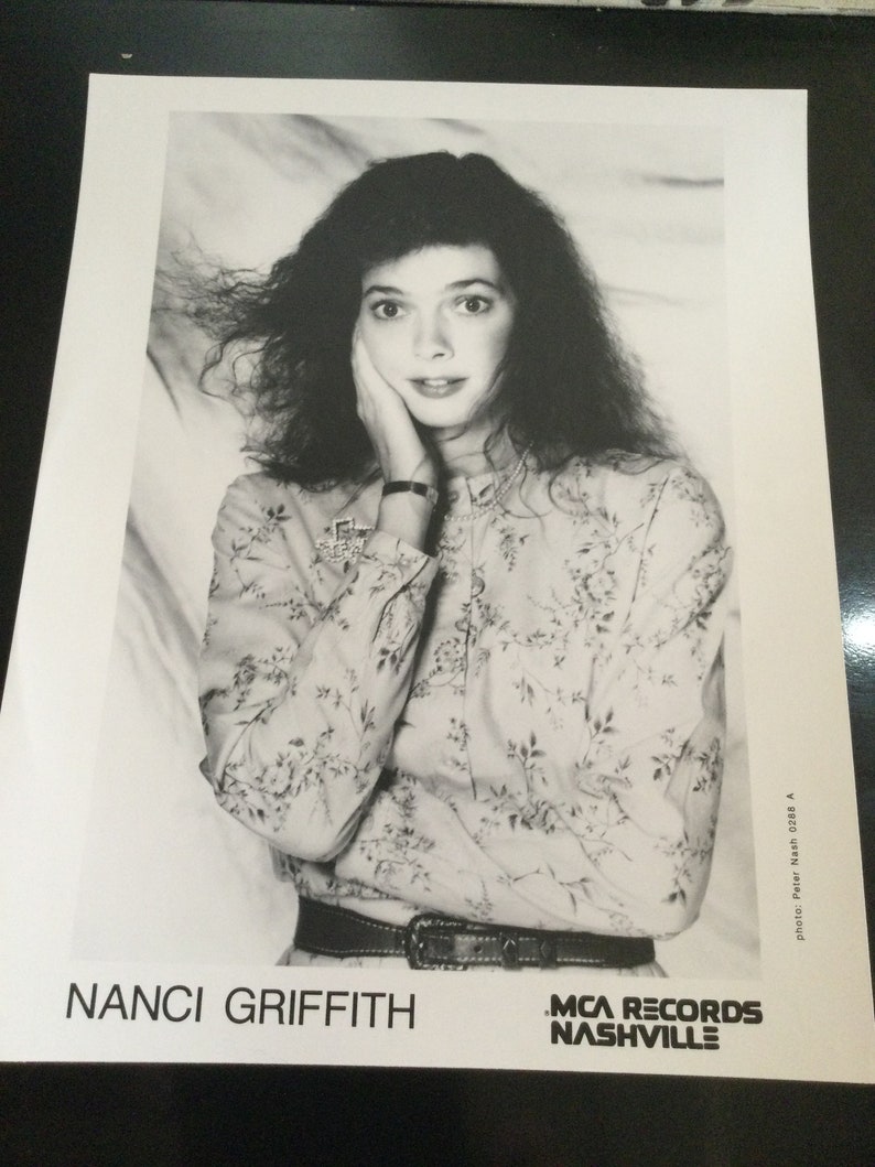 May include: Black and white promotional photo of Nanci Griffith. She is wearing a floral print blouse and a belt. Her dark hair is styled with volume. The photo includes the text "NANCI GRIFFITH" and "MCA RECORDS NASHVILLE".