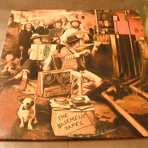 May include: Album cover for "The Basement Tapes" featuring a sepia-toned image of a band playing instruments in a basement setting. A dog sits in the foreground. The album title is handwritten on a box.