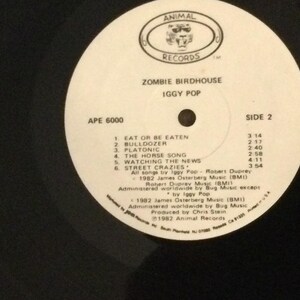 May include: A white vinyl record labeled "ZOMBIE BIRDHOUSE" by Iggy Pop. The record features a track list for Side 2, including songs like "Eat or Be Eaten" and "Watching the News." The record label includes the "ANIMAL RECORDS" logo.