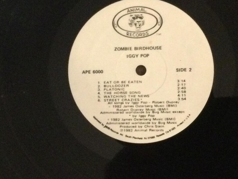 May include: A white vinyl record labeled "ZOMBIE BIRDHOUSE" by Iggy Pop. The record features a track list for Side 2, including songs like "Eat or Be Eaten" and "Watching the News." The record label includes the "ANIMAL RECORDS" logo.