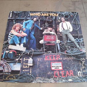 May include: Album cover for "Who Are You" by The Who. The cover features the band members posing in front of a backdrop of electrical cables and equipment. The album title is at the top, with the words "NOTICE ELECTRICAL AREA CLEAR" visible.