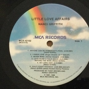 May include: A vinyl record for the album "Little Love Affairs" by Nanci Griffith. The record label features a rainbow over a cloudy sky. The album title and artist name are in white text. The label also lists song titles and credits.