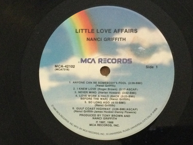 May include: A vinyl record for the album "Little Love Affairs" by Nanci Griffith. The record label features a rainbow over a cloudy sky. The album title and artist name are in white text. The label also lists song titles and credits.