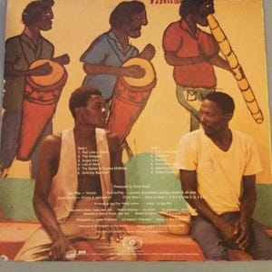 May include: Album cover featuring a colorful illustration of musicians playing drums and a wind instrument. Two men are seated in the foreground. The album title and track list are visible.