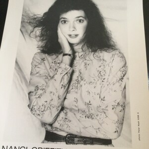 May include: Black and white photograph of Nanci Griffith, with long, dark, curly hair. She wears a floral print blouse, a belt, and a watch. The image includes the text "NANCI GRIFFITH" and "MCA RECORDS NASHVILLE."